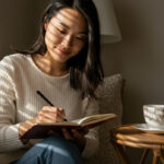 Woman smiling while writing in journal at home with coffee cup, documenting family legacy stories and memories